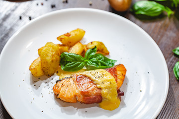 Grilled salmon fillet wrapped in bacon and potato wedges on white plate. Close up