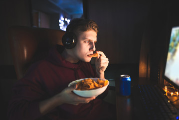 Young man eats chips at night, drinks beer and uses a computer. Portrait of a gamer playing video games on a computer and eating snacks. Supper at the computer at night. © bodnarphoto