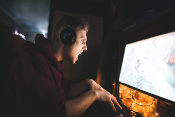 Expressive young gamer plays games at home coma in a cozy room at home. Portrait of an evil gamer in headphones playing video games on a computer. - obrazy, fototapety, plakaty