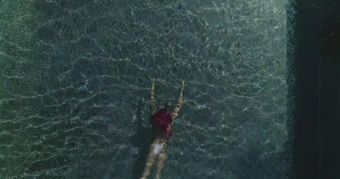 AERIAL CLOSE UP TOP DOWN: Young woman on summer vacation jumping head first into clear blue pool water for refreshment on hot sunny day. Girl in pink bikini diving into empty pool in luxury resort