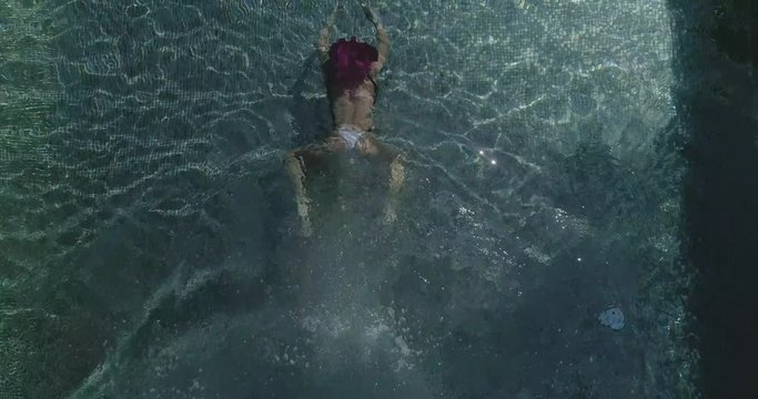 AERIAL CLOSE UP TOP DOWN: Young woman on summer vacation jumping head first into clear blue pool water for refreshment on hot sunny day. Girl in pink bikini diving into empty pool in luxury resort