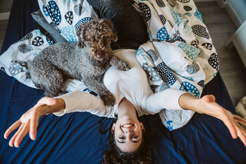 .Young and funny woman playing with her nice spanish water dog on top of the bed, freshly awake on a winter morning. Lifestyle.