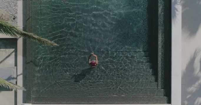 AERIAL CLOSE UP TOP DOWN: Young woman on summer vacation jumping head first into clear blue pool water for refreshment on hot sunny day. Girl in pink bikini diving into empty pool in luxury resort