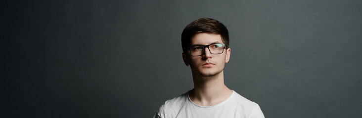 Portrait of pensive teen guy dressed in white, wearing glasses and standing near black background....