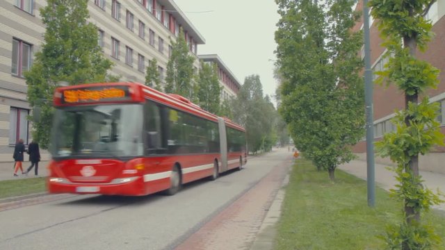 A long red bus driving down a narrow tree lined street between modern concrete buildings on a college campus. People walk on sidewalks beside a busy road. Student housing and dormitories.