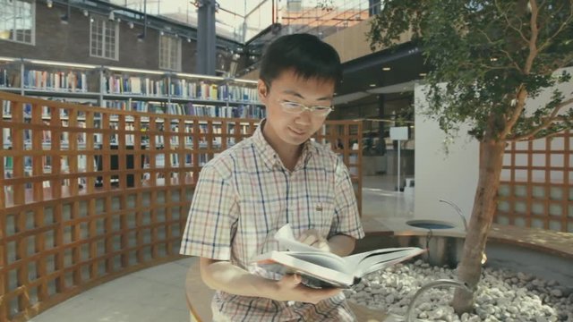 A young Asian man reading a book in the university library. Man with glasses looking at a book. Chinese student sits on a wall around a tree planted inside a building. TRACKING.