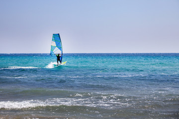 Surfer Spending Time on Outdoor Water Sport Adventure. Clear Sky and Blue Wave. Person Balancing on a Windsurfing Board with Sail. Vacation Activity ideas