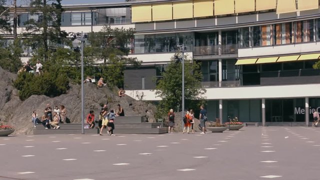 Wide plaza with plenty of benches and large rocks for people to sit on and round cement planter boxes filled with flowers in front of a large modern office building. People outside on a sunny day.