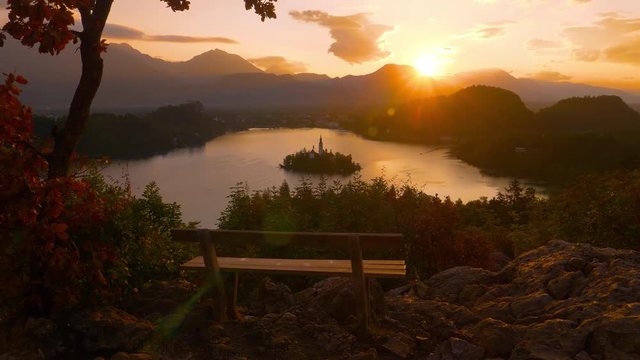 CLOSE UP LENS FLARE: Breathtaking shot of sunset illuminating empty bench overlooking beautiful lake Bled and the small island. Scenic shot of a wooden bench above the famous lake on sunny morning.