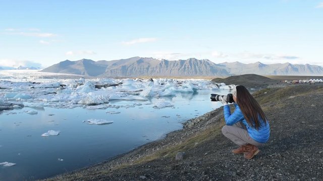 Photographer tourist woman taking photos with DSLR camera on travel on Iceland by Jokulsarlon glacial lagoon / glacier lake on Iceland. Happy tourist girl on travel in beautiful nature landscape.