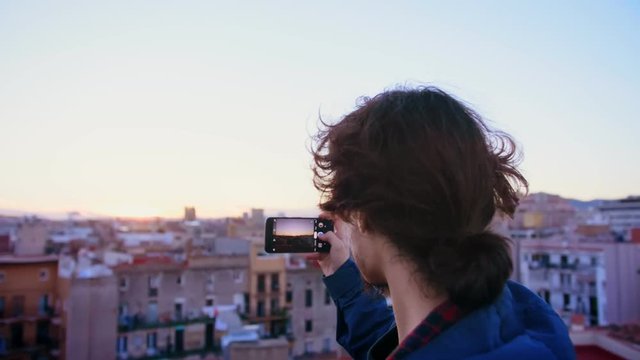 Young hipster student in Erasmus new in city learning photography as a hobby and making smartphone or mobile phone camera photo or picture of view of rooftop during sunset or sunrise in Europe town