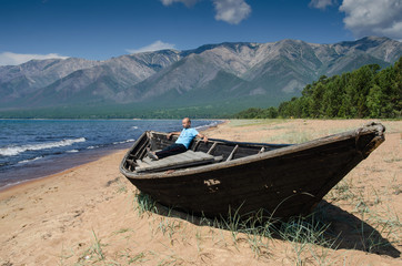 Men is sitting in very old wooden fishing boat aground on the beach, near lake Baikal, Russia, on a sunny day in the summer
