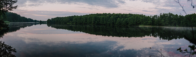 Lake with spring trees panorama photo. Tranquil landscape at a lake, with the vibrant blue sky, white clouds and the trees reflected symmetrically in the clean blue water.