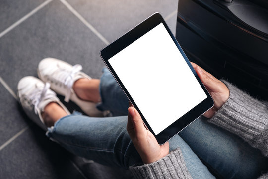 Top view mockup image of woman's hands holding and using black tablet pc with blank white desktop screen while sitting on the floor