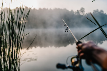 Fisherman with fishing rod, spinning reel on the background river bank. Sunrise. Fog against the...