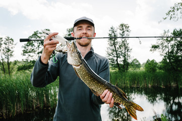 Happy cheerful young fisherman hold a big fish pike on bridge, pier on a background of lake and...
