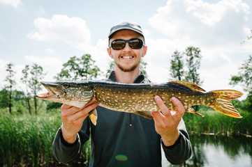 Happy cheerful young fisherman hold a big fish pike on a background of lake and nature. Fishing...