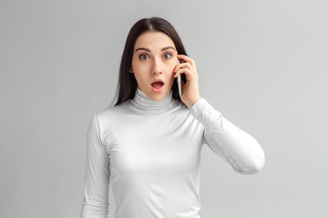 Freestyle. Woman standing isolated on gray talking answering phone call looking camera shocked