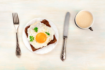 A plate with an egg cooked sunny side up on a toast with coffee, a fork, and a knife, shot from the top with copy space