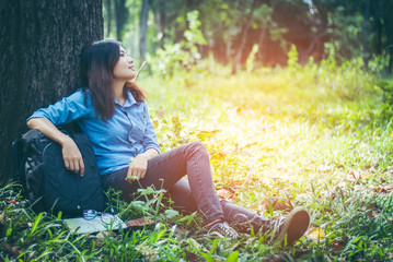 In tropical forests, hiking Asian girl with a backpack resting under the shade of large trees. She leaned against a tree and smiling happily on holiday weekends with copy space.