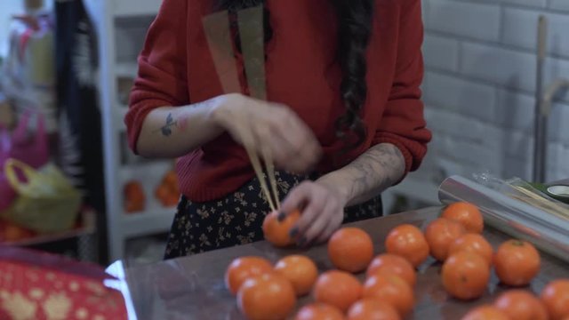 Woman is piercing through tangerines with sharp wooden stick and putting them together. Delicious fruit-floral atmosphere
