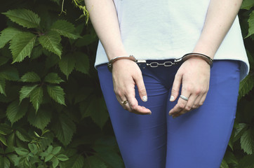 Fragment of a young criminal girl's body with hands in handcuffs against a green blossoming ivy...