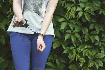 Fragment of a young criminal girl's body with hands in handcuffs against a green blossoming ivy...