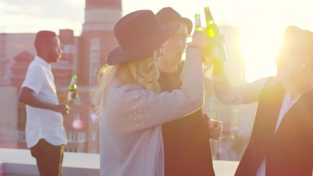 Young hipster friends in fedora hats toasting with beer and dancing while having fun at sunset party on urban rooftop