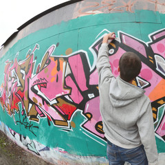 A young guy in a gray hoodie paints graffiti in pink and green c