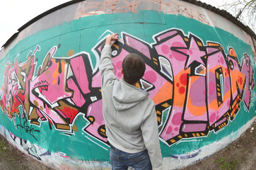 A young guy in a gray hoodie paints graffiti in pink and green c