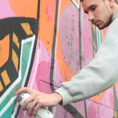 A young guy in a gray hoodie paints graffiti in pink and green c