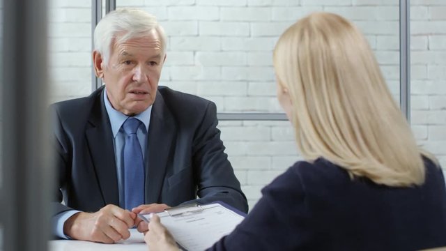 Tracking shot of senior man in suit and necktie giving CV on clipboard to female manager with blond hair and talking about his skills and experience during job interview