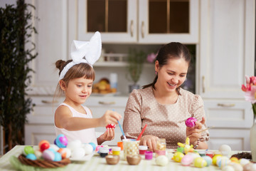 Happy young mother and her two little daughters with white rabbit's ears on their heads dye the eggs for the Easter table in the cozy light kitchen