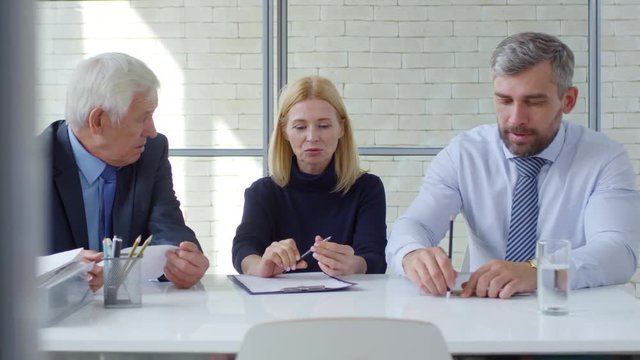 Tracking shot of elderly businessman in suit, blond businesswoman and bearded manager wearing necktie sitting at table and discussing job candidate after panel interview