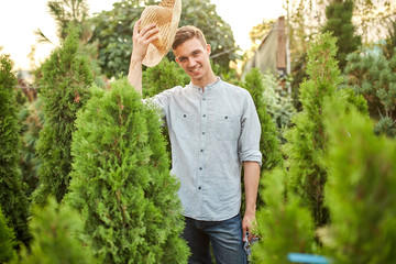 Smiling guy gardener in a straw hat stands in the nursery-garden with a lot of thujas on a warm sunny day