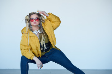 young blonde girl in sunglasses with african pigtails listens to music in headphones and dances against a white background.