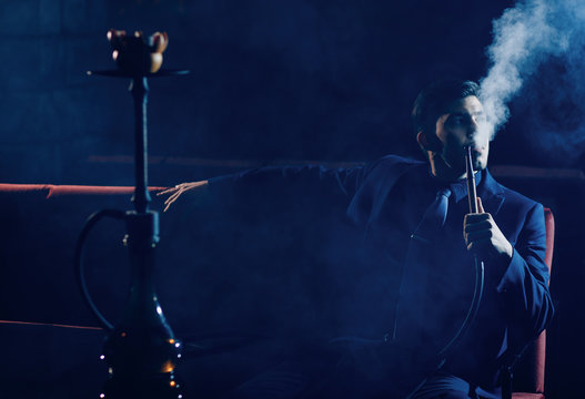 Smoking hookah, young man is resting in nightclub, dark background, white cloud of smoke