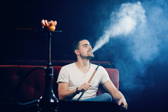 Smoking hookah, young man is resting in nightclub, dark background, white cloud of smoke