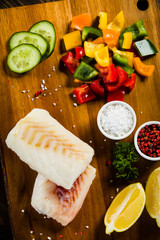 Fresh raw cod with herbs and vegetables served on cutting board on wooden table