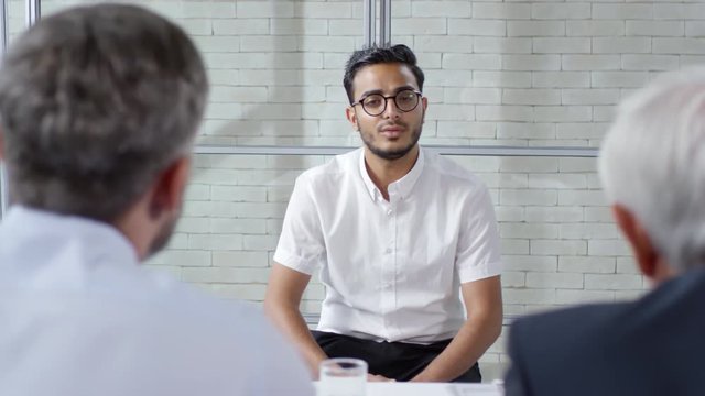 Tracking shot of young Arab man in formalwear sitting opposite elderly businessman and bearded manager conducting job interview. He is talking about himself and smiling