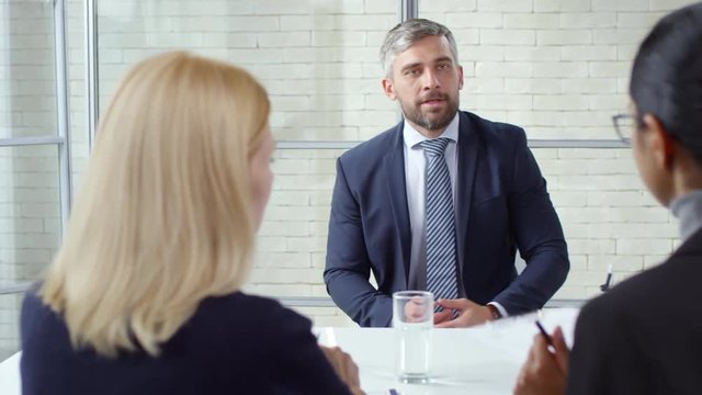 Tracking shot of bearded man in suit and necktie giving his CV to businesswomen during panel job interview and talking about his professional experience