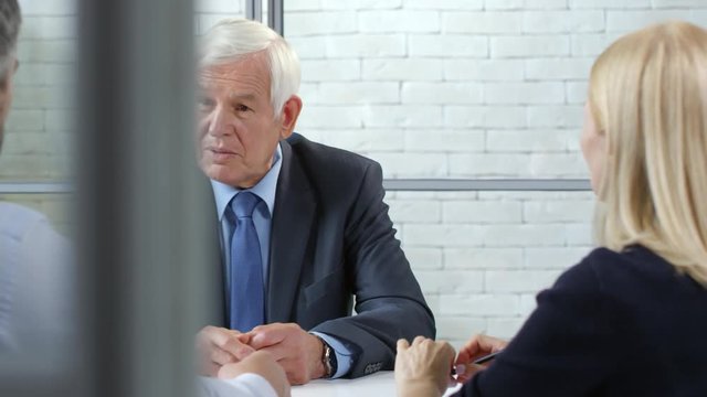 Tracking shot of elderly male executive in suit and necktie sitting opposite businessman and businesswoman and gesturing while discussing work
