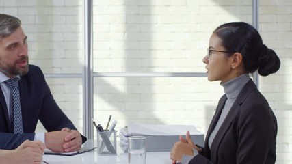 Tracking shot of confident young woman in glasses and formalwear talking to businessman with beard and blond businesswoman during job interview