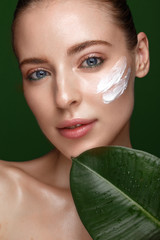 Beautiful fresh girl with cosmetic cream on the face, natural make-up and green leaves. Beauty face.