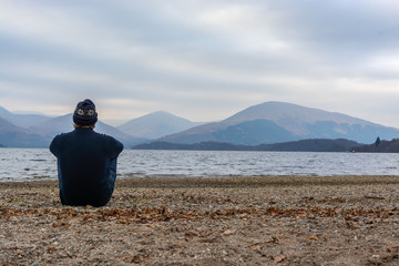 Man in solitude sitting on a messy beach. Background is beautiful mountain range in scotland. Taken at Long Lomond during winter.