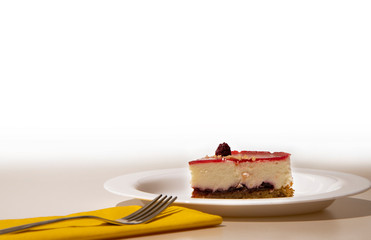 a slice of cheesecake on a white plate, with yellow napkin and steel cake fork. White background.