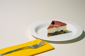 a slice of cheesecake on a white plate, with yellow napkin and steel cake fork. White background.