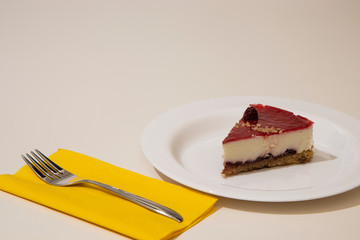 a slice of cheesecake on a white plate, with yellow napkin and steel cake fork. White background.