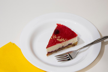 a slice of cheesecake on a white plate, with yellow napkin and steel cake fork. White background.