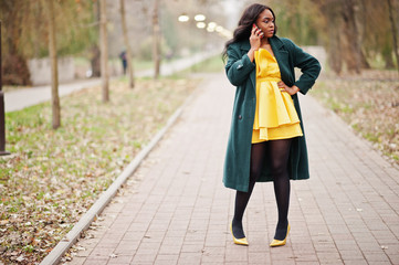 Stylish african american woman at green coat and yellow dress posed against autumn park and speaking on phone.